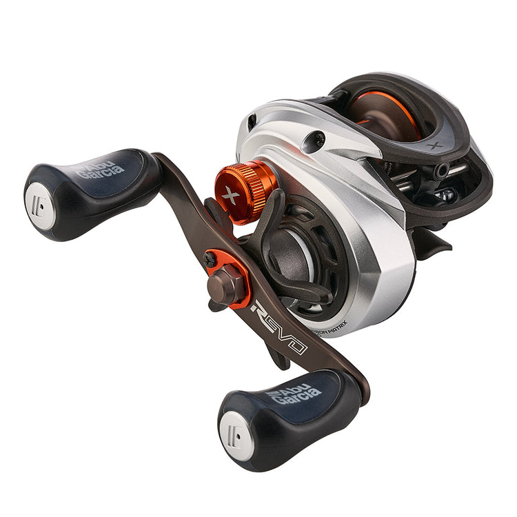 A close-up of a silver and black baitcasting fishing reel with orange accents, featuring two handles and visible gears. The reel is marked with brand symbols on the handle grips.