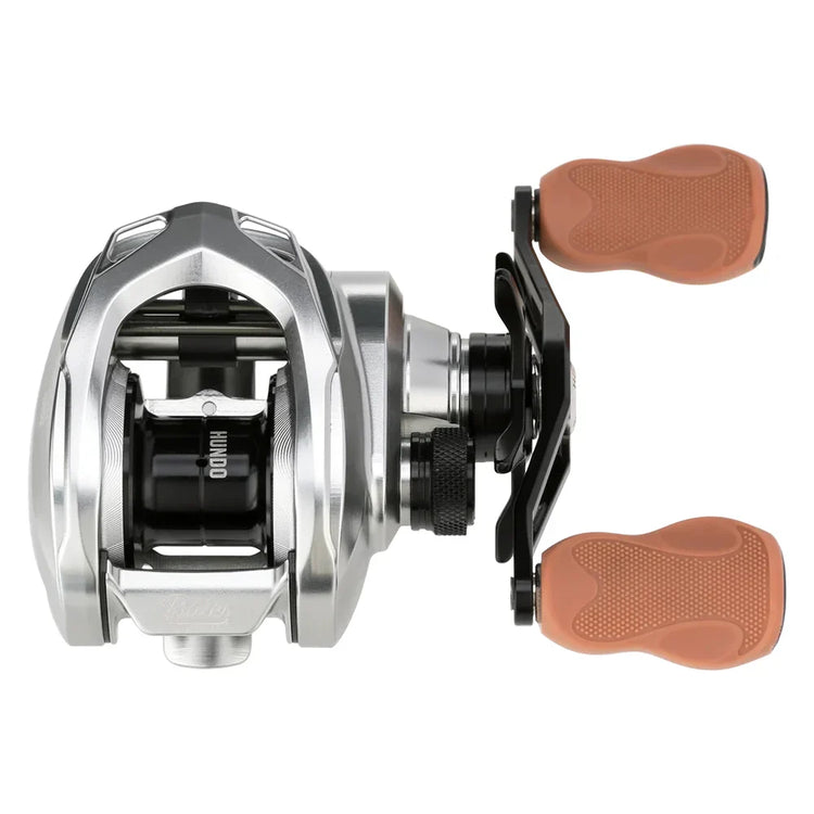 A silver baitcasting fishing reel with dual brown handles is pictured against a white background. The reel features a compact design and a visible spool mechanism.