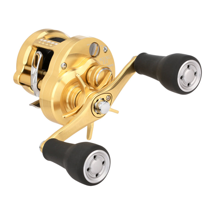 A gold fishing reel with black handles, showing a side view. The reel features intricate metalwork and a shiny finish, highlighting its design. The two crank handles are positioned on the right side.