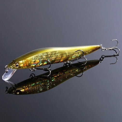 The glossy gold Megabass Vision 110 Jr. Jerkbait with reflective scales and three treble hooks lies on a shiny black surface. Featuring a clear curved lip, it's designed to mimic small fish, luring predators with the innovative Multiway Moving Balancer System.