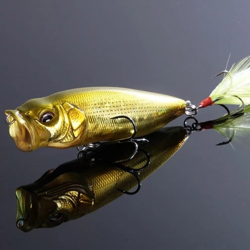 The Megabass PopMax, a gold-colored fishing lure from Megabass with realistic fish details, rests on a reflective black surface. It features a feathered tail hook and advanced gill system, designed intricately for baiting fish as an effective topwater lure.