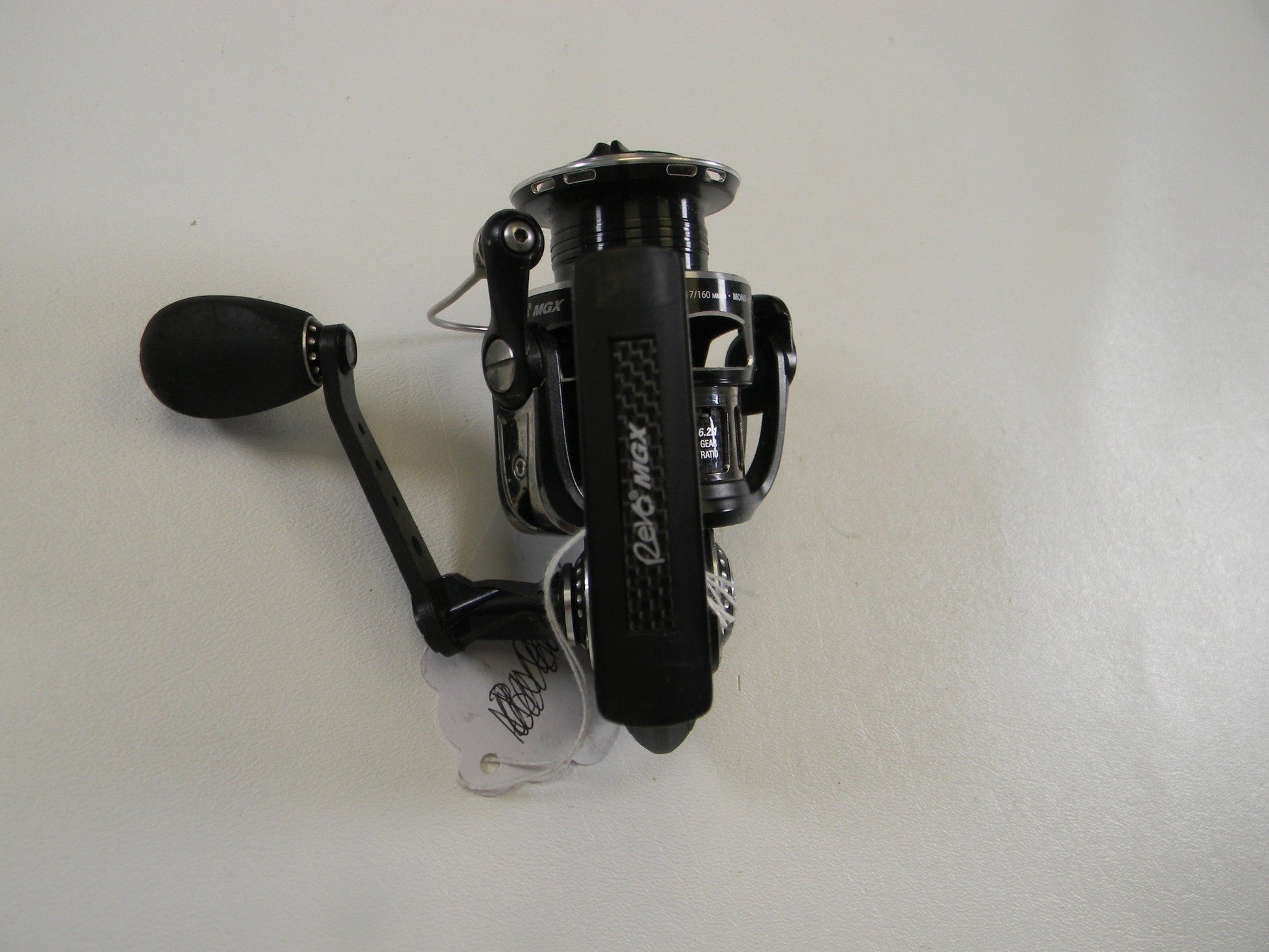 On a white background, the Abu Garcia Revo MGX 30 6.2:1 - Used Spinning Reel in black with a crank handle and branded label is displayed, connected to a price tag. Ideal for those participating in Rod and Reel Trade-In or looking to earn store credit by exchanging used gear.