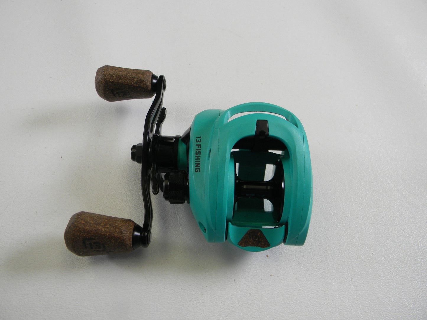 The 13 Fishing Concept TX2 TX2-7.5-LH, a teal reel with a black frame and cork-like brown handles, sits on a white background, ready for your next trade-in opportunity.