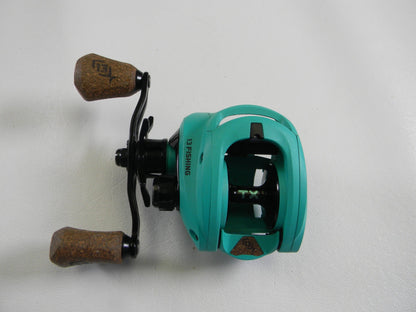 A teal 13 Fishing Concept TX2 casting reel with brown cork handles, black metal components, and the "13 FISHING" logo rests on a white surface. Ideal for those considering used gear or involved in a Rod and Reel Trade-In for store credit.