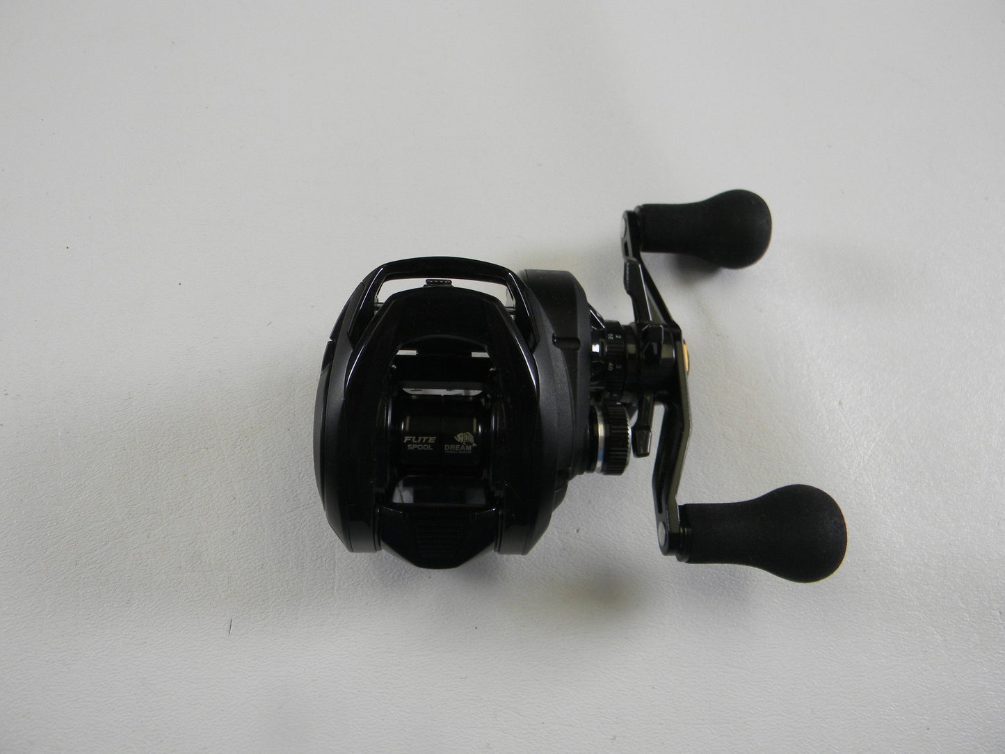 The Okuma Hakai HDT 100H 7.3:1 RH, a black baitcasting reel with two handles in excellent condition, is on a white surface—ideal for upgrading your used gear through our Rod and Reel Trade-In program.