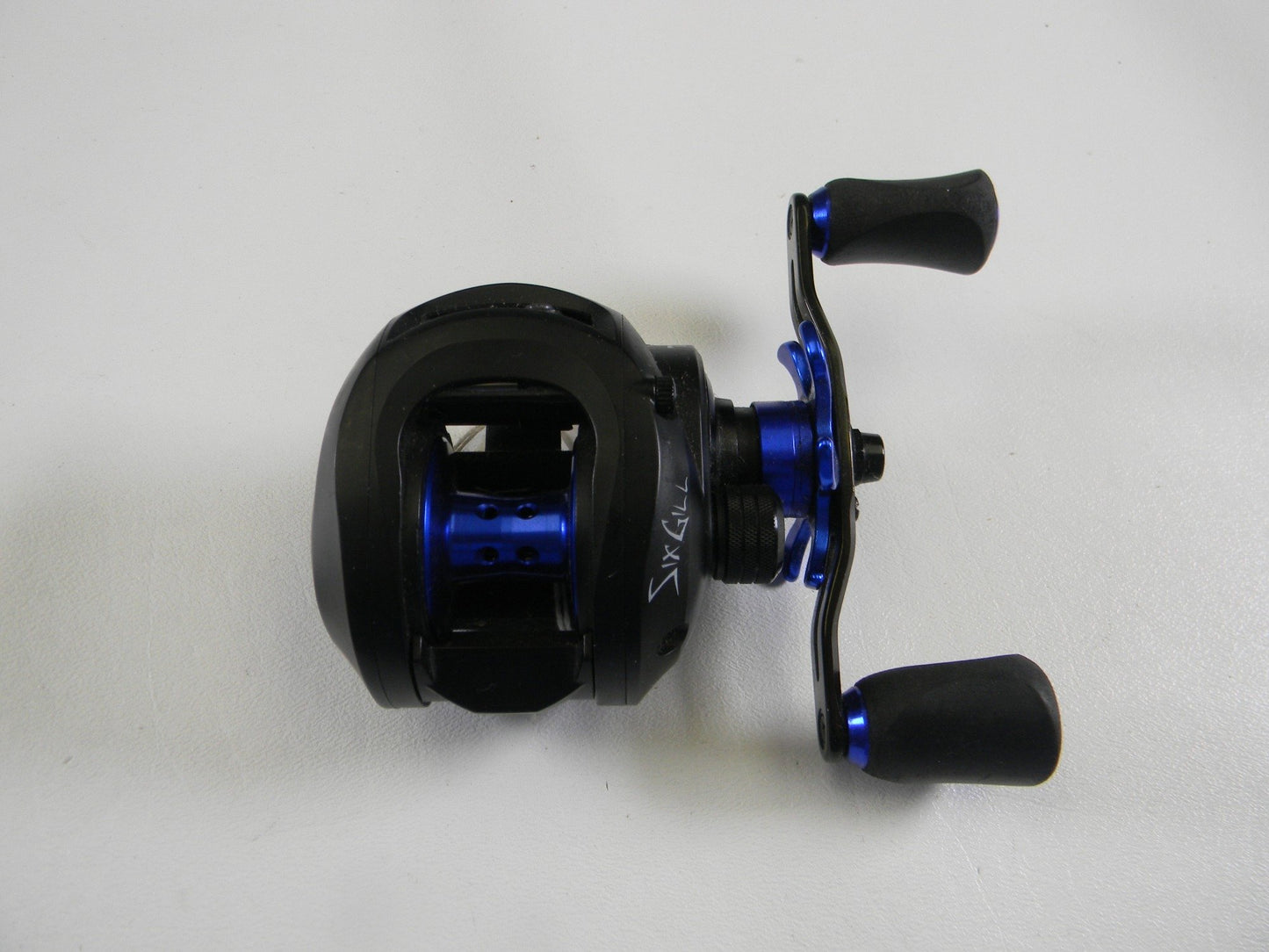 This used SixGill Cyclone 7.1:1 RH baitcasting reel in black and blue, featuring dual handles and stylish visible markings, is available for Rod and Reel Trade-In. It is in good condition and showcased against a white background.