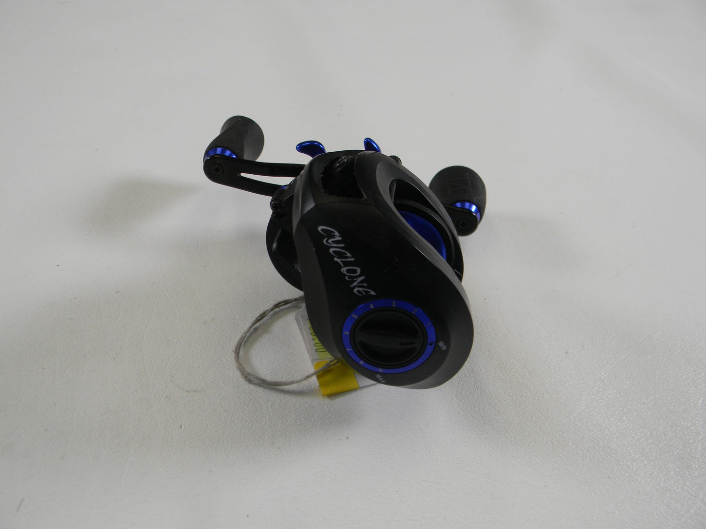 A SixGill Cyclone 7.1:1 RH used casting reel in good condition, featuring a black and blue design with dual grips, lies on a white surface with a yellow tag. It's part of the Rod and Reel Trade-In, ideal for earning store credit.