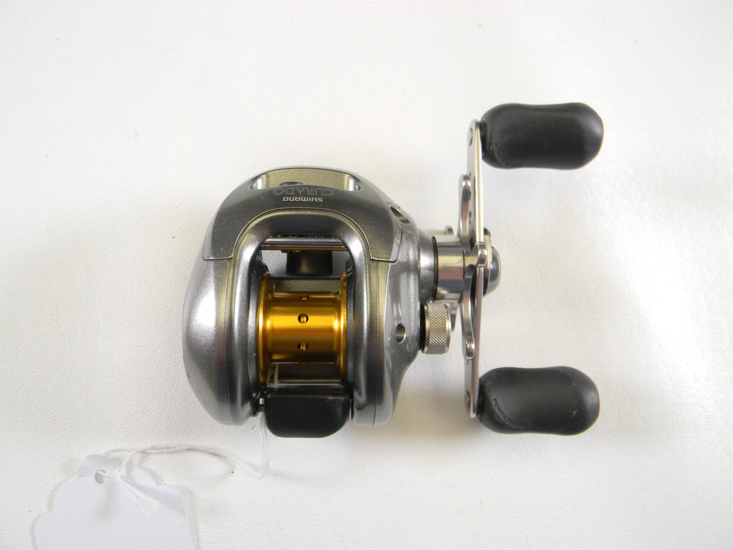 A Shimano Curado 100D CU100D, a silver baitcasting reel with a gold spool, dual-handle grip, and small white tag, shines against a plain backdrop. Ideal for anglers looking to upgrade through our Rod and Reel Trade-In events for store credit on quality used gear.