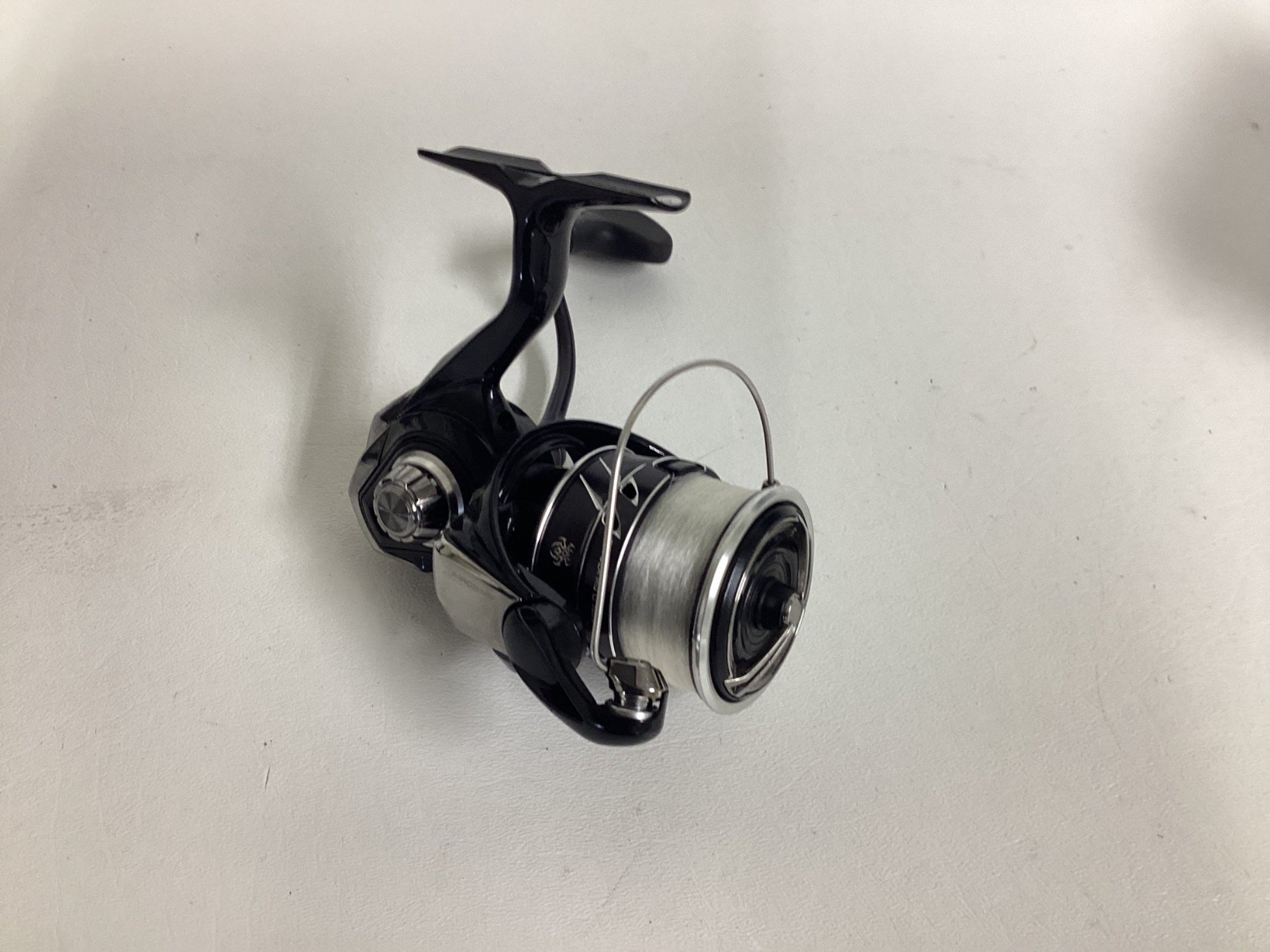 A black and silver Daiwa Tatula MQ LT3000-CXH fishing reel sits on a white surface, ready for adventure. Its sleek design includes a spool filled with line and a winding handle. Trade in your used gear through the Rod and Reel Trade-In Program for store credit toward your next upgrade.