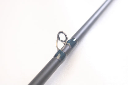 A close-up of a St. Croix Physyx PHXC76MHMF 7'6" Medium Heavy casting rod guide features a metallic ring on the glossy, dark rod. Part of our exclusive Rod and Reel Trade-In Program, it offers great value for your used gear. The softly blurred background highlights the guide's secure wrap.