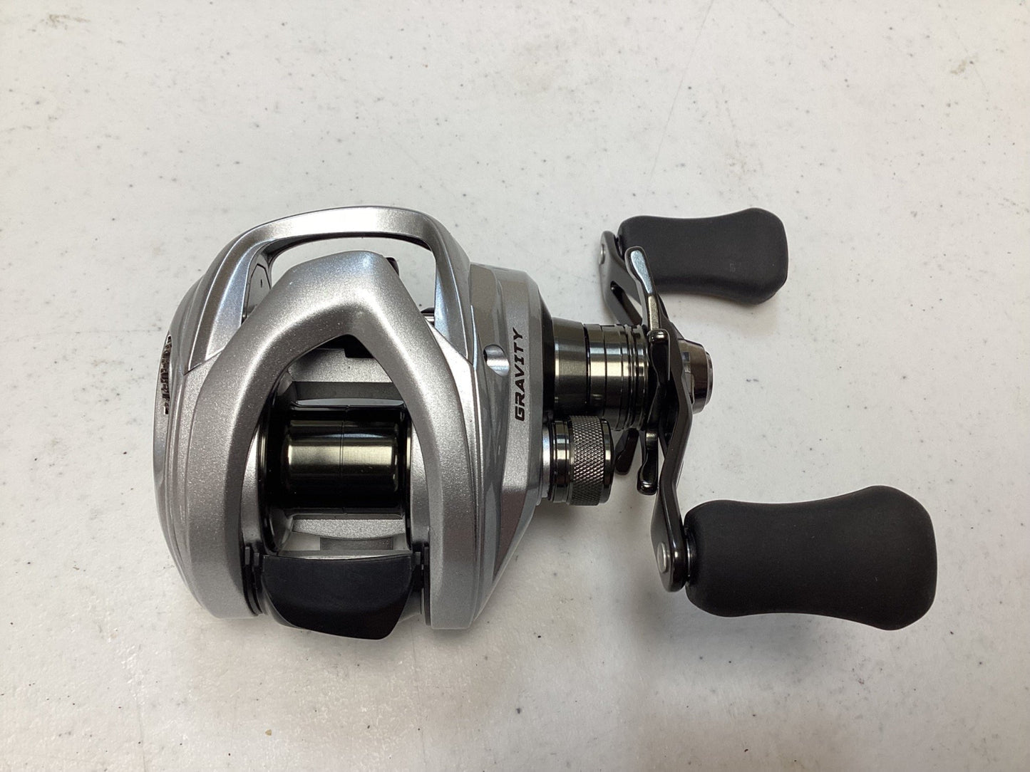 A sleek silver casting reel, Ark Gravity 7 G7107 7.1:1 RH in excellent condition, featuring a black handle and grip, rests on a white surface. Ideal for upgrading used gear with our Rod and Reel Trade-In program for store credit.