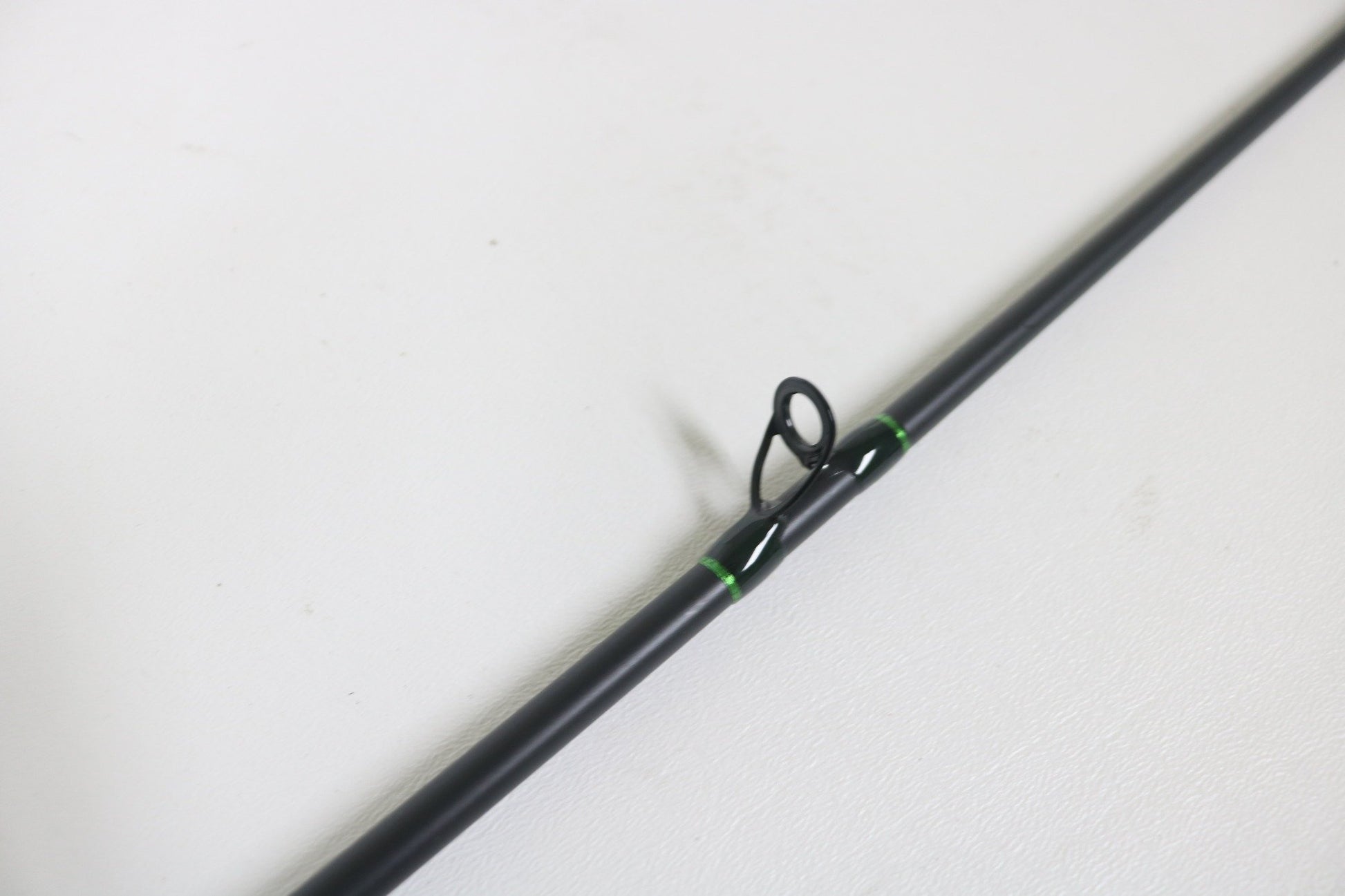 Close-up of a G. Loomis IMX-Pro 722C CR Used Casting Rod in mint condition against a light gray background. The sleek black rod features a single circular eyelet reinforced with green wraps, ideal for those upgrading through our Rod and Reel Trade-In program for store credit.