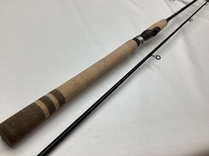 A G. Loomis IMX-PRO Salmon/Steelhead Float 1260-2S STFR 10'6" light spinning rod with a cork handle and black body lies on a white surface, split into two pieces to display the guides and reel seat. Mint condition.