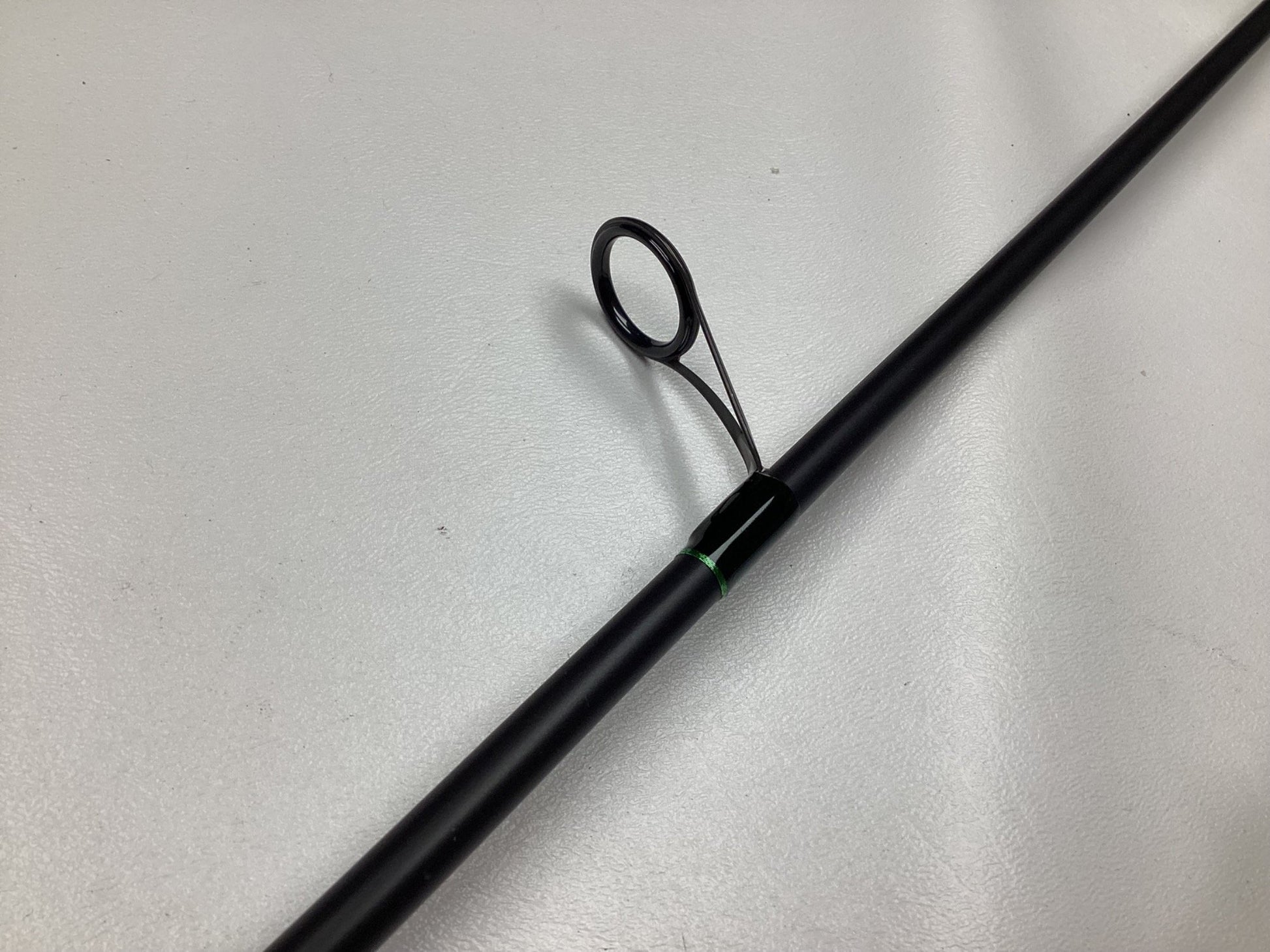 Close-up of a G. Loomis IMX-PRO black fishing rod with a green accent, laying on a light gray surface, featuring one prominent circular guide for the fishing line.