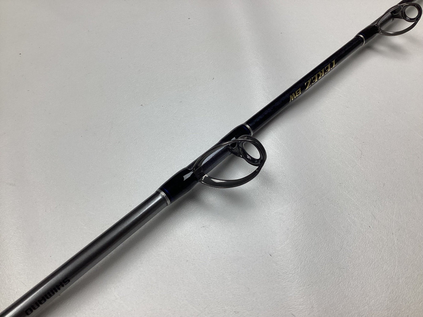 Close-up of a used Shimano Terez BW Conventional Slick Butt Rod 6'6" Medium Heavy in mint condition, lying on a light surface. The black rod has circular guides and gold text, ideal for those joining our rod and reel trade-in program or earning store credit.