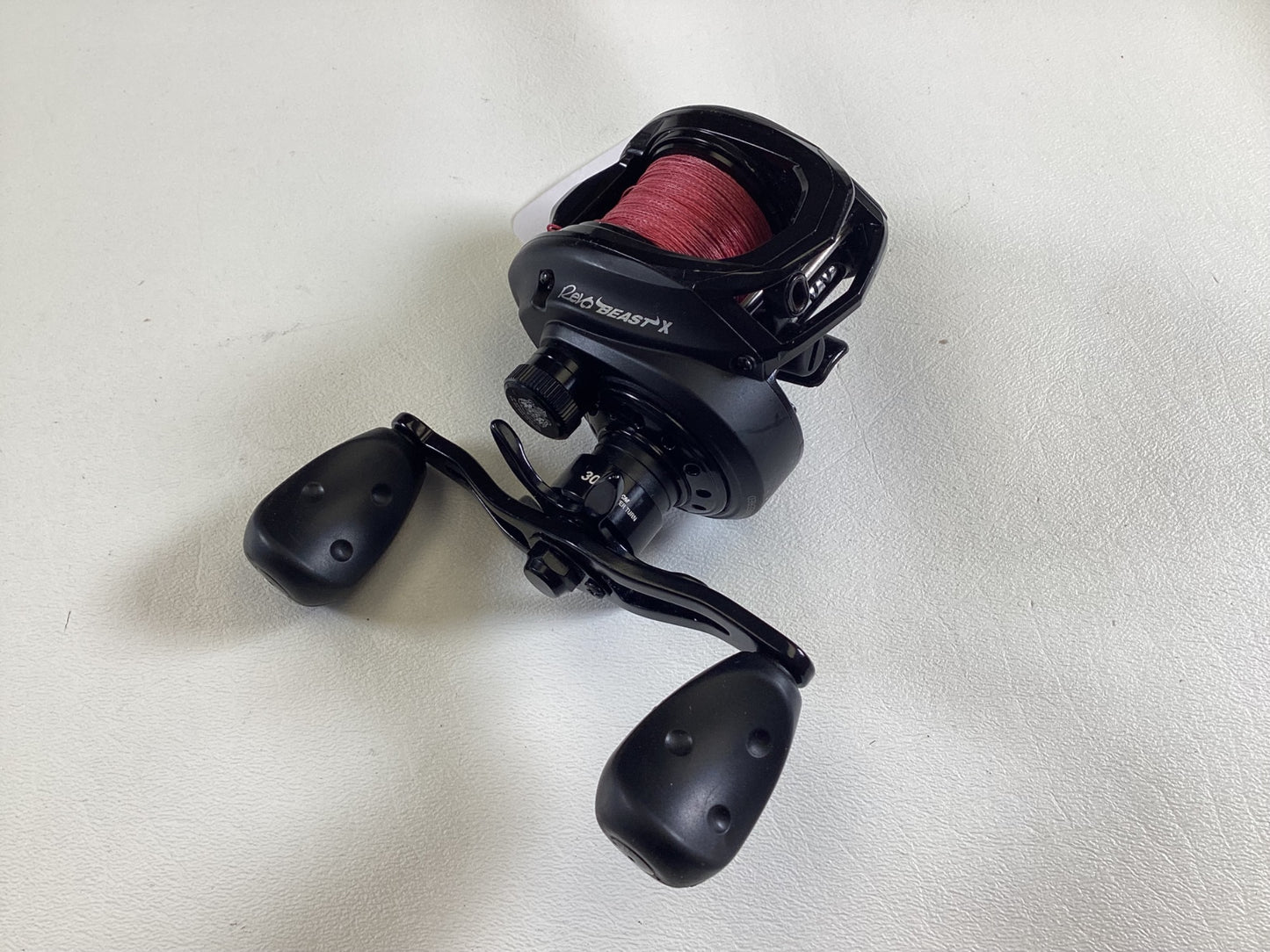 A black Abu Garcia Revo Beast X 40 reel with red line rests on a white surface, featuring large handle knobs. Ideal for anglers ready to upgrade through our Rod and Reel Trade-In program.