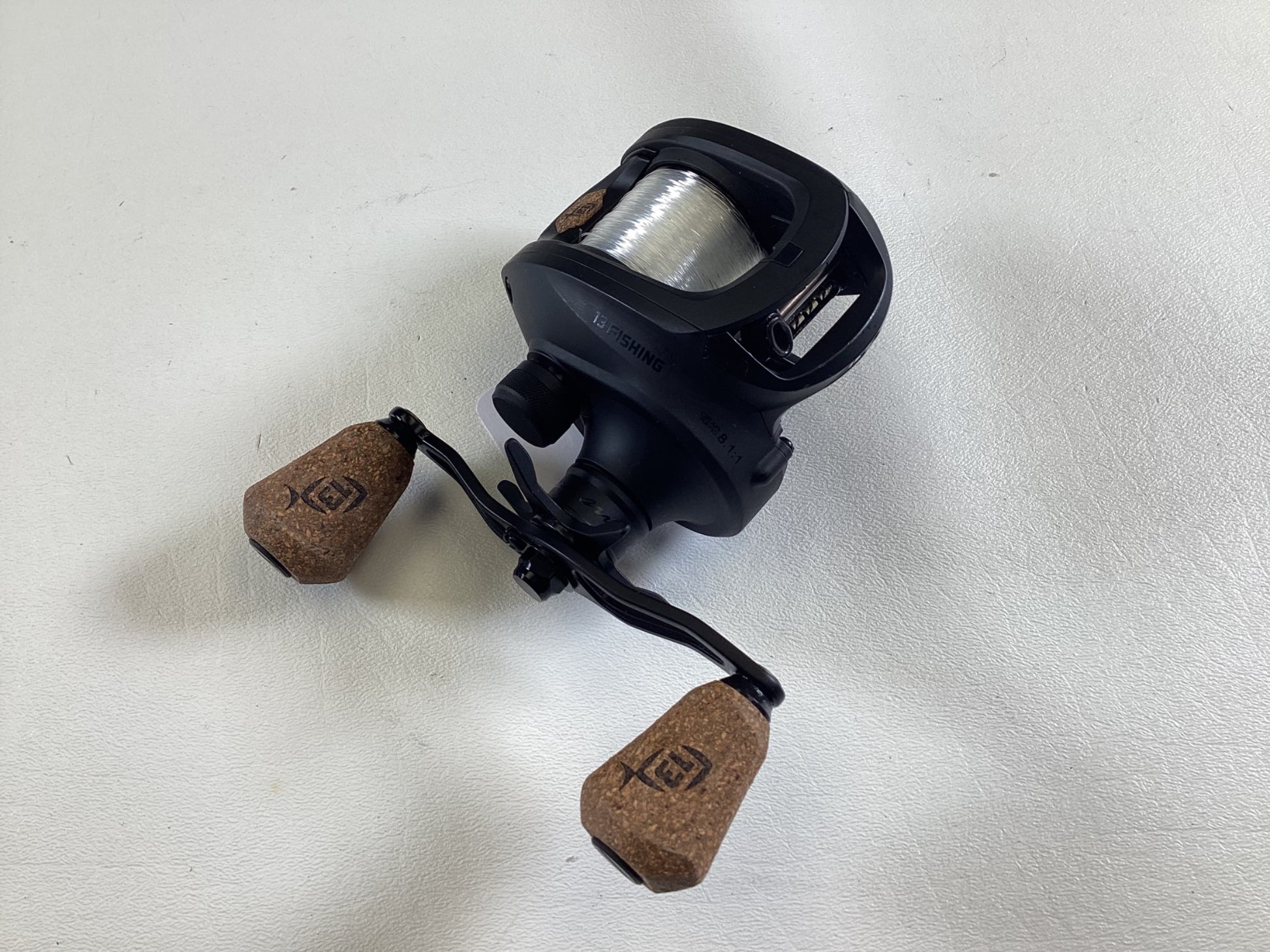 The 13 Fishing Concept A3 Gen II casting reel, with cork handles and partially wound fishing line, gleams on a white background showcasing the brand logo. Ideal for the Rod and Reel Trade-In program, this black reel is rated in excellent condition.