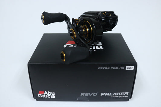 A close-up of a black Abu Garcia Revo Premier Gen 4 casting reel with gold accents, model "REVO4 PRM-HS" and gear ratio 7.3:1, resting on its box. This used gear is perfect for our Rod and Reel Trade-In Program, offering you an opportunity to earn store credit!.