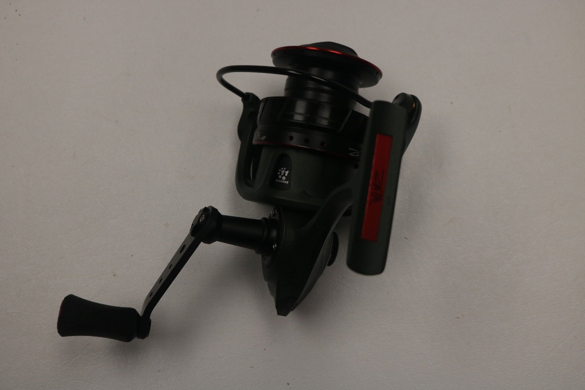 The Abu Garcia Zata ZATASP40 6.2:1, a used spinning reel in excellent condition, features a winding handle and line spool in black and green with red accents on a white surface. Ideal for anglers, upgrade your gear through our Rod and Reel Trade-In Program.