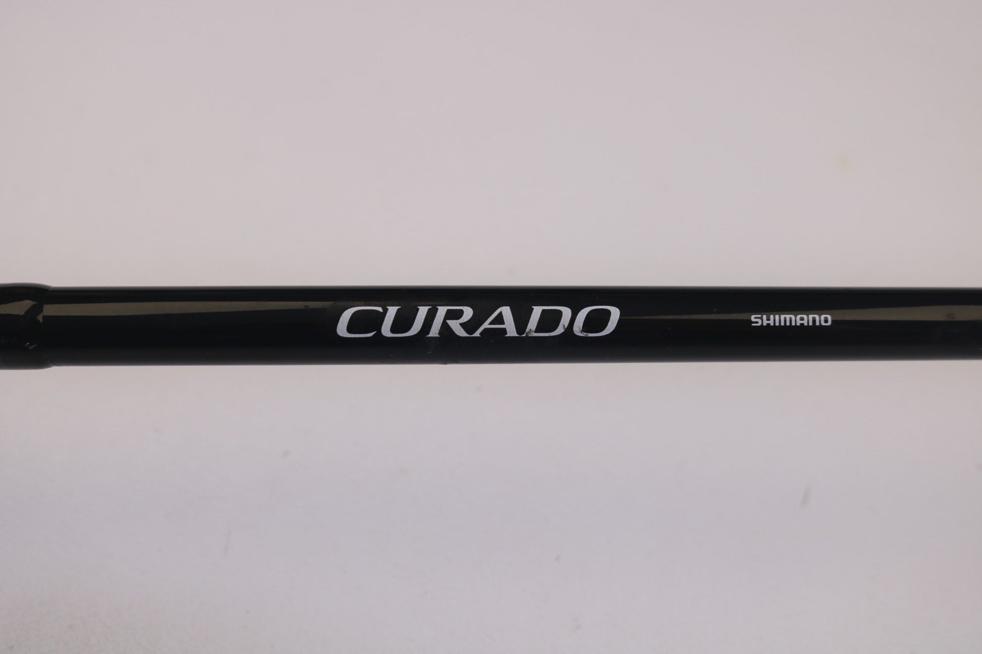 Close-up image of a Shimano Curado CDC610M 6'10" Medium, with "CURADO" in large and "SHIMANO" in small font on its dark surface. Ideal for our Rod and Reel Trade-In Program, where used gear can be exchanged for store credit. Background is plain white.