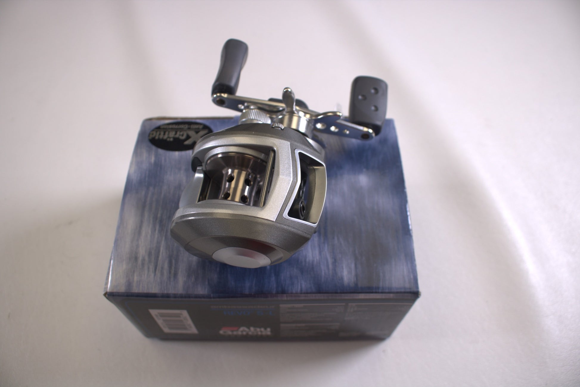 An Abu Garcia Revo S-L Gen 1 6.4:1 LH used casting reel in excellent condition with a black handle sits on a blue and black box on a white surface. This metallic reel, featuring some transparent parts, is eligible for our Rod and Reel Trade-In Program, offering store credit for your used gear.