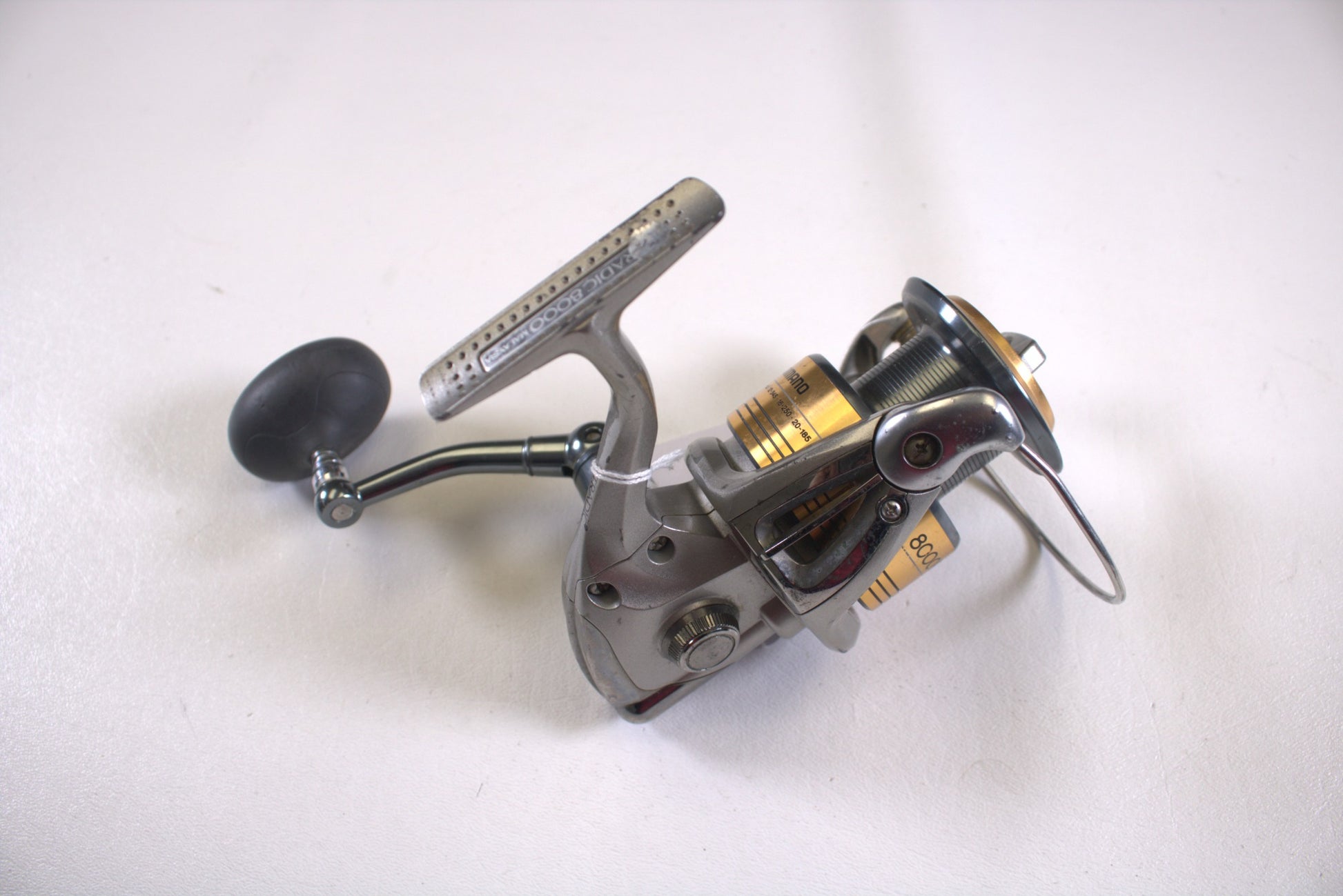 The Shimano Stradic ST8000FI, a used spinning reel in fair condition with missing line keeper, features a silver body with a black handle and gold accents on a white surface. Trade in your gear as part of our Rod and Reel Trade-In Program for potential store credit today.