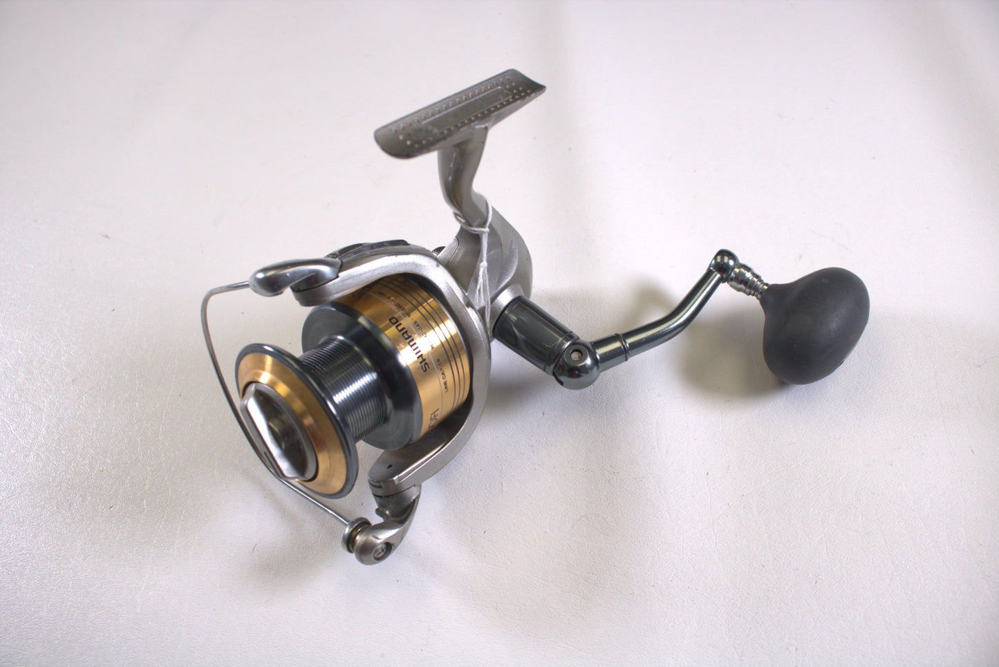 The Shimano Stradic ST8000FI used spinning reel in silver and gold, featuring a black handle, rests on a white surface. Its durable metal arm and spool make it ideal for the Rod and Reel Trade-In Program, while its sleek round grip blends style with functionality. Condition: Fair, missing line keeper.