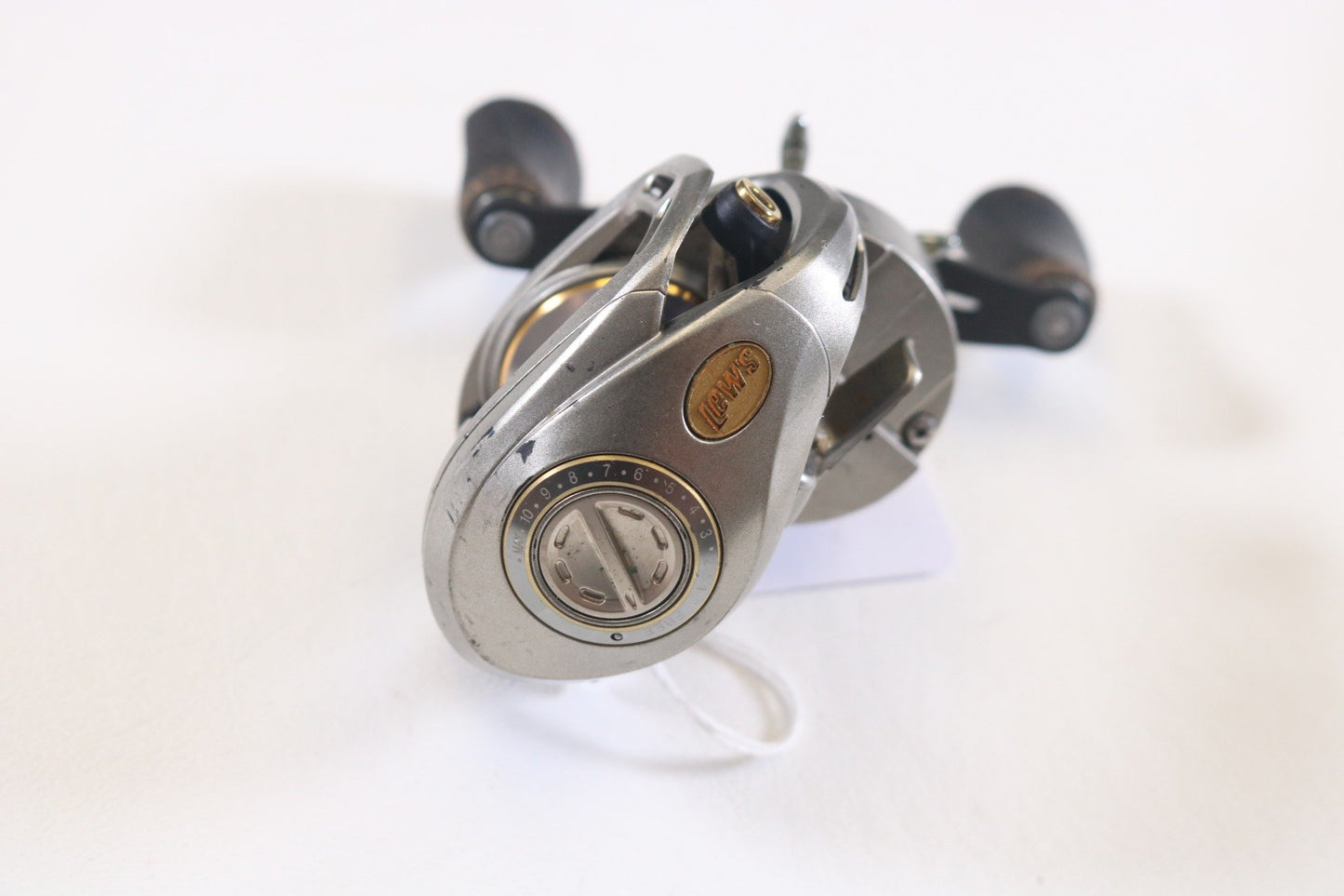 A close-up of the Lew's Team Lew’s Lite TLL1SHL 7.5:1 LH casting reel in good condition shows a sleek, modern design, ideal for the Rod and Reel Trade-In Program. It has two handles with dark knobs and a small side dial, resting on a white surface with a slight shadow.